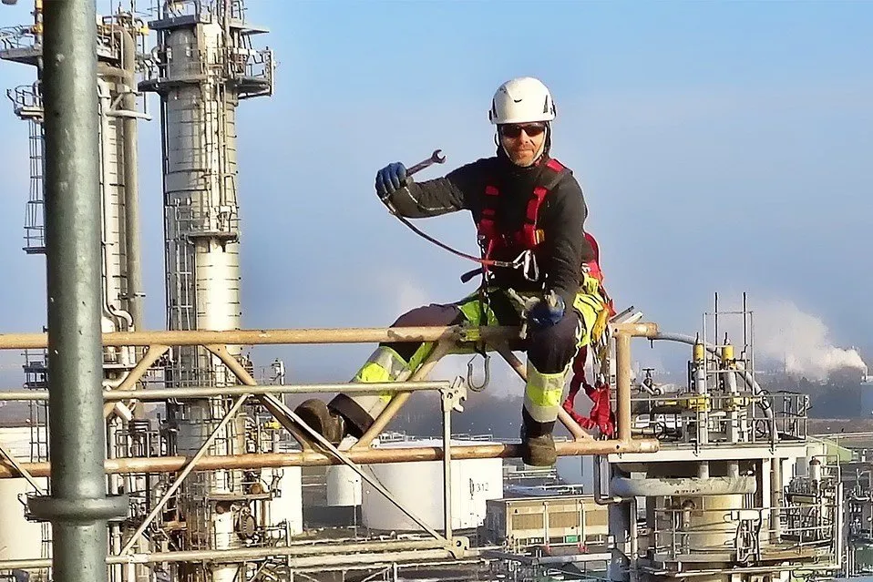En man vinkar till kameran med ett verktyg i handen samtidigt som han arbetar med sanering via industriklättring.